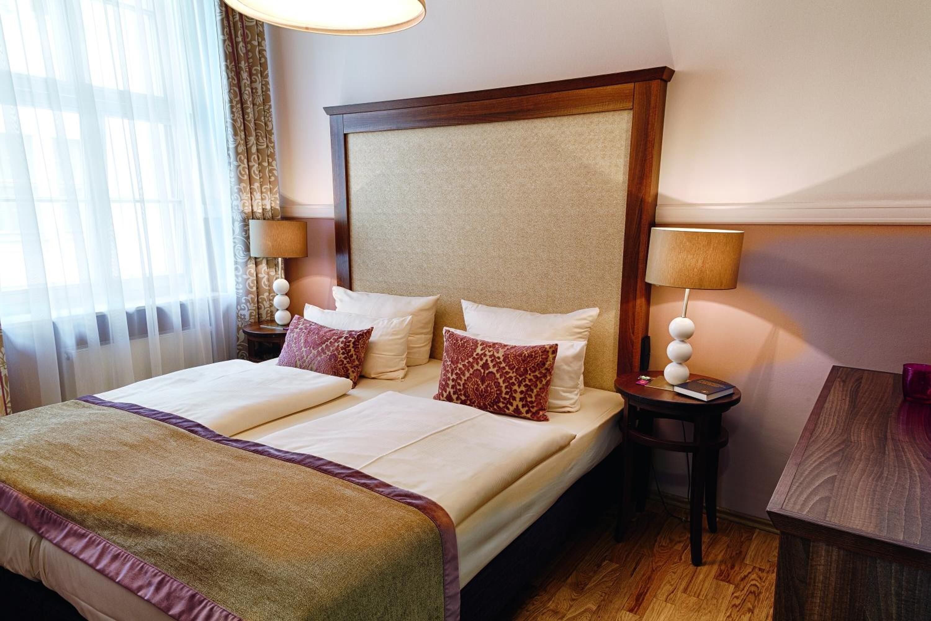 A hotel room with a large bed, two bedside tables, a small table, and a window with curtains.