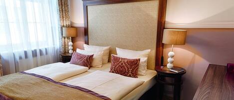 A hotel room with a large bed, two bedside tables, a small table, and a window with curtains.