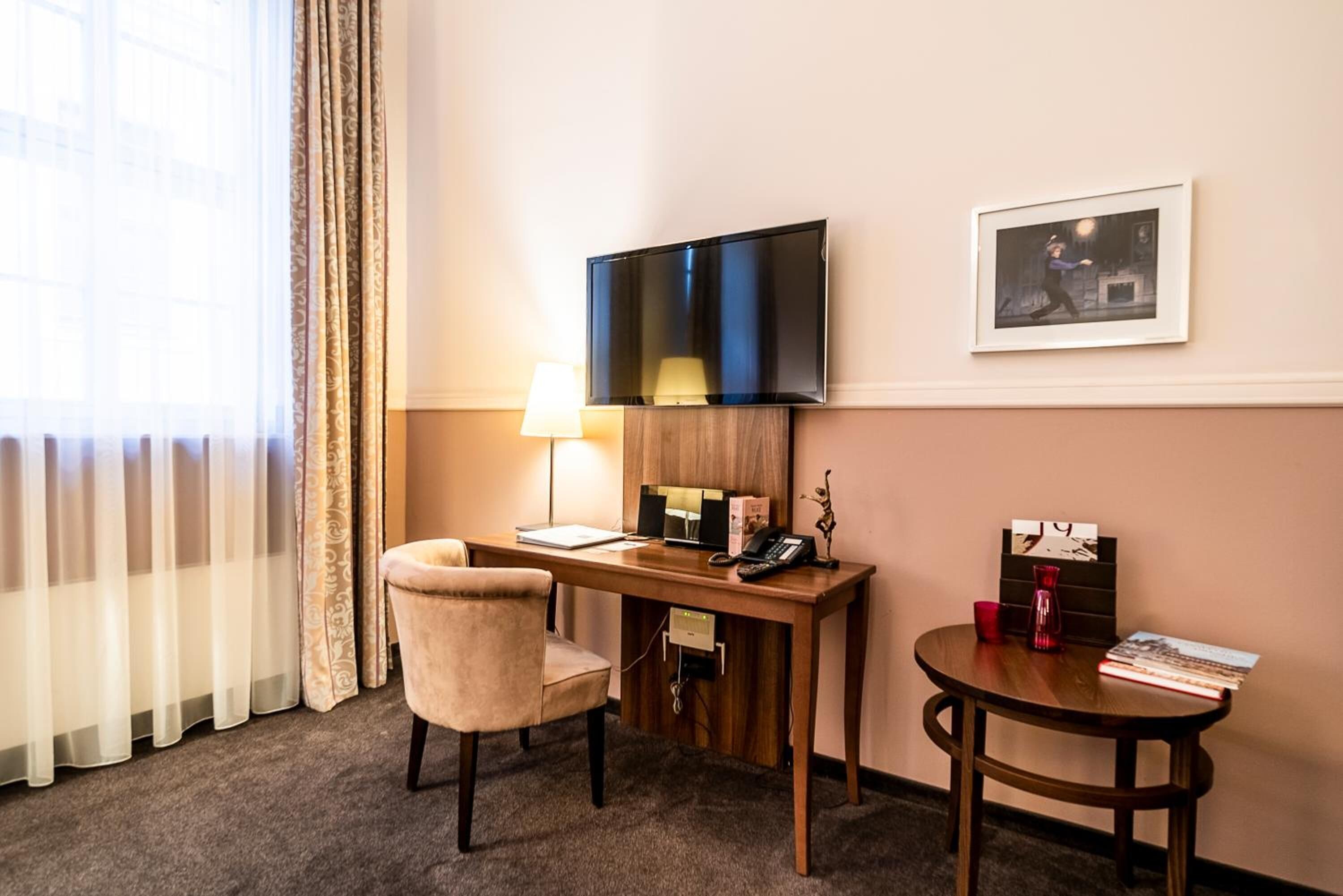 A hotel room with a desk, chair, TV, lamp, framed picture, and a small table with books and a vase.