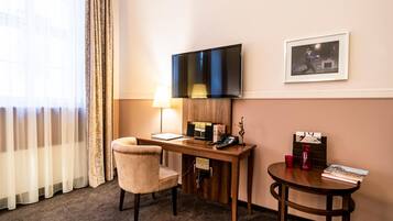 A hotel room with a desk, chair, TV, lamp, framed picture, and a small table with books and a vase.