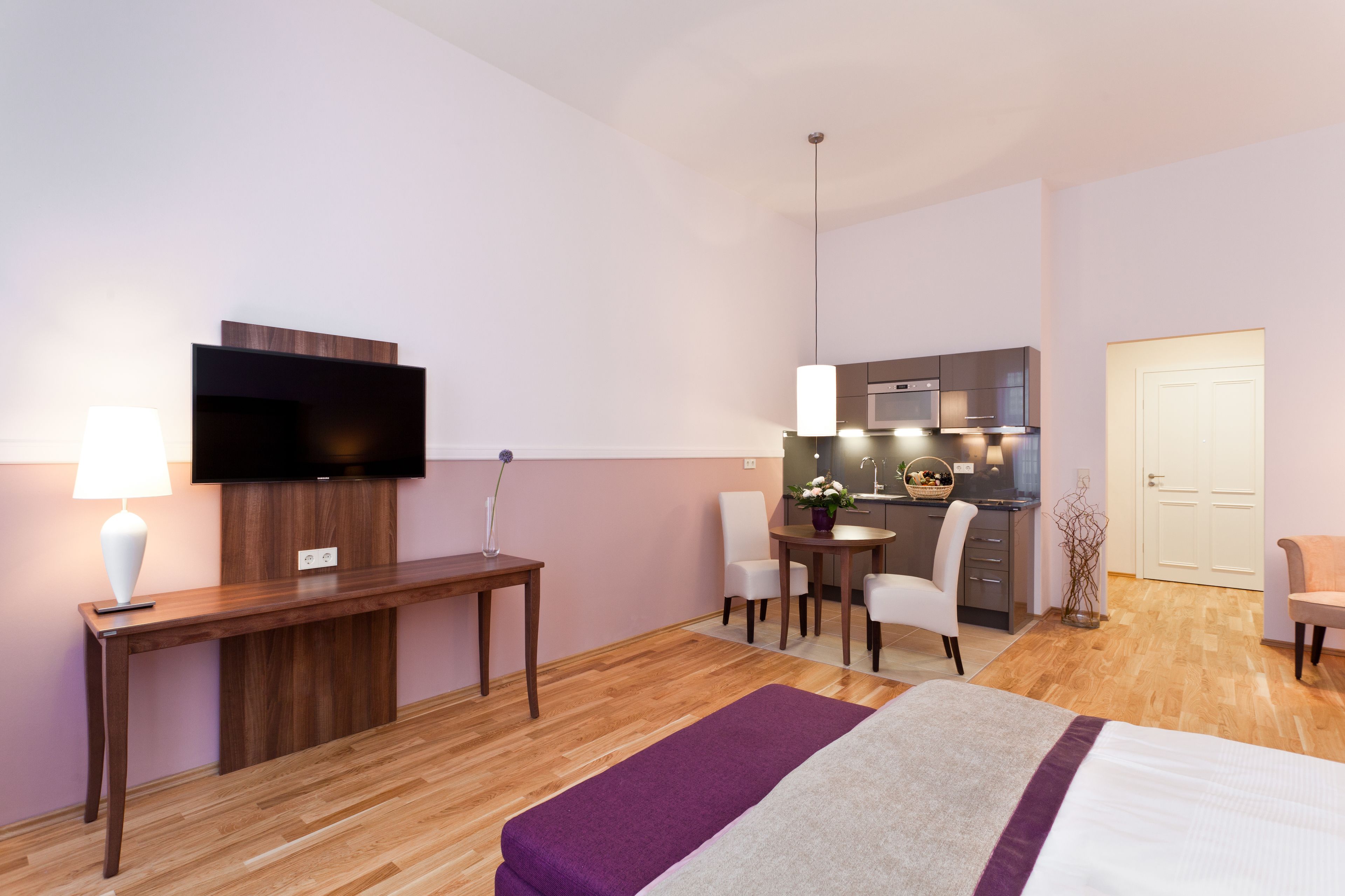 A modern hotel room with a wooden desk, a flat-screen TV, a dining area with a table and chairs, a kitchenette, and a bed with a purple and white bedspread.