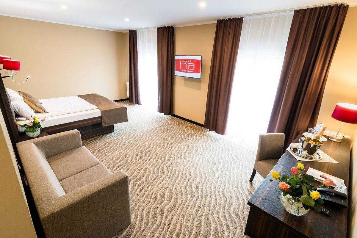 Habitación de hotel con cama, sofá, escritorio con lámpara, florero con flores y un cartel en la pared.