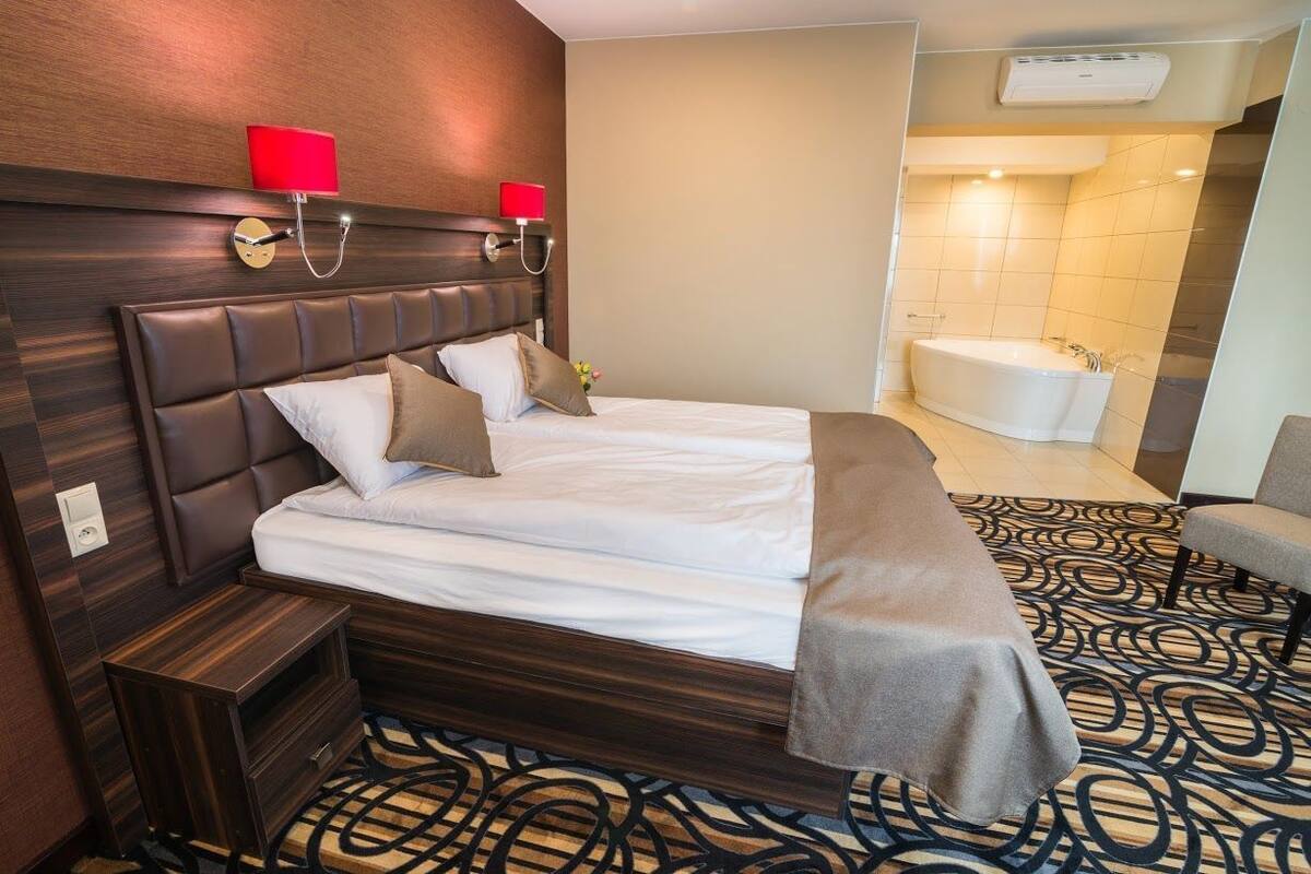Habitación de hotel con una cama grande, una mesita de noche, una lámpara roja y un baño con bañera y ducha.