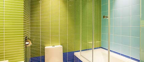 Une salle de bain avec des murs recouverts de carreaux verts et bleus, des toilettes blanches et une douche en verre.