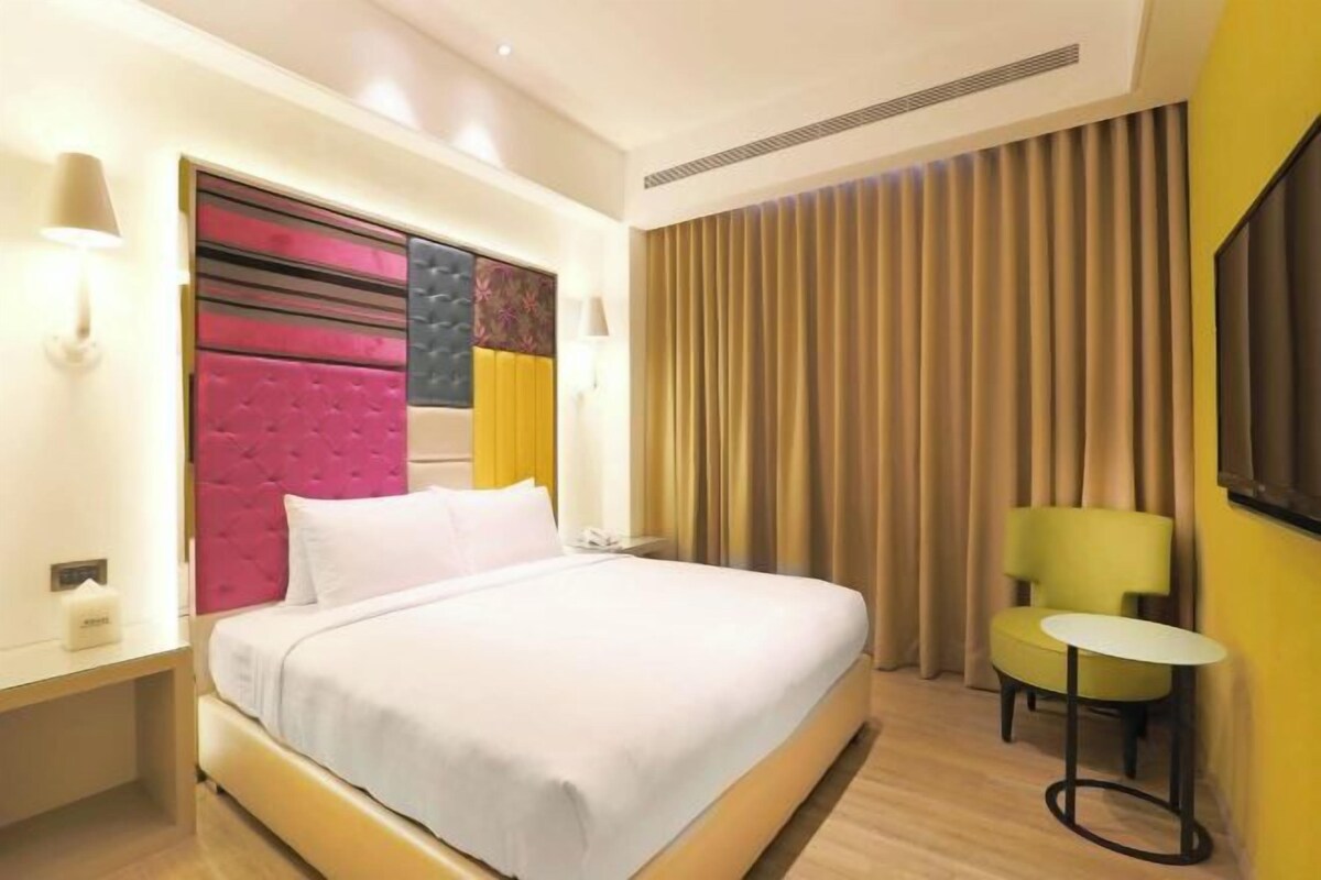 Una habitación de hotel con una cama grande, cabecera con paneles rosa y negro, una mesita y una silla.