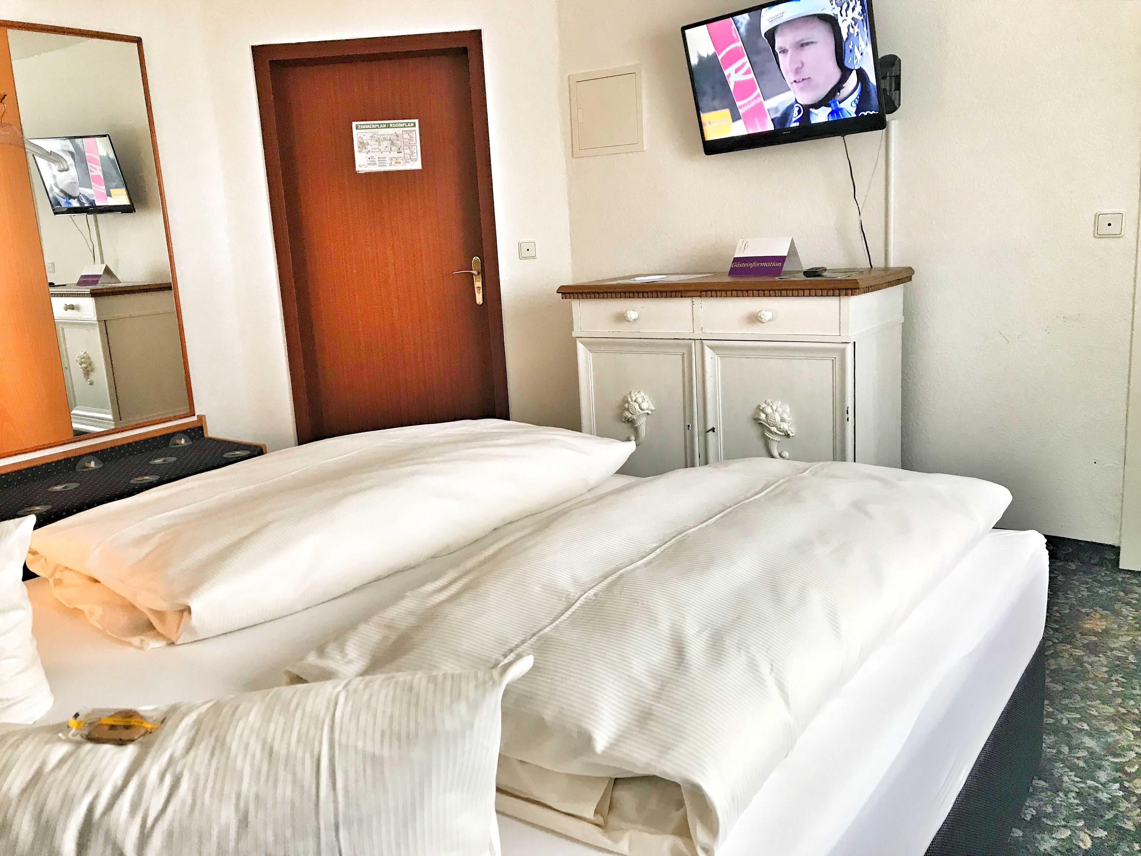 Une chambre d’hôtel avec deux lits, une porte en bois, un téléviseur fixé au mur et une commode avec un miroir.