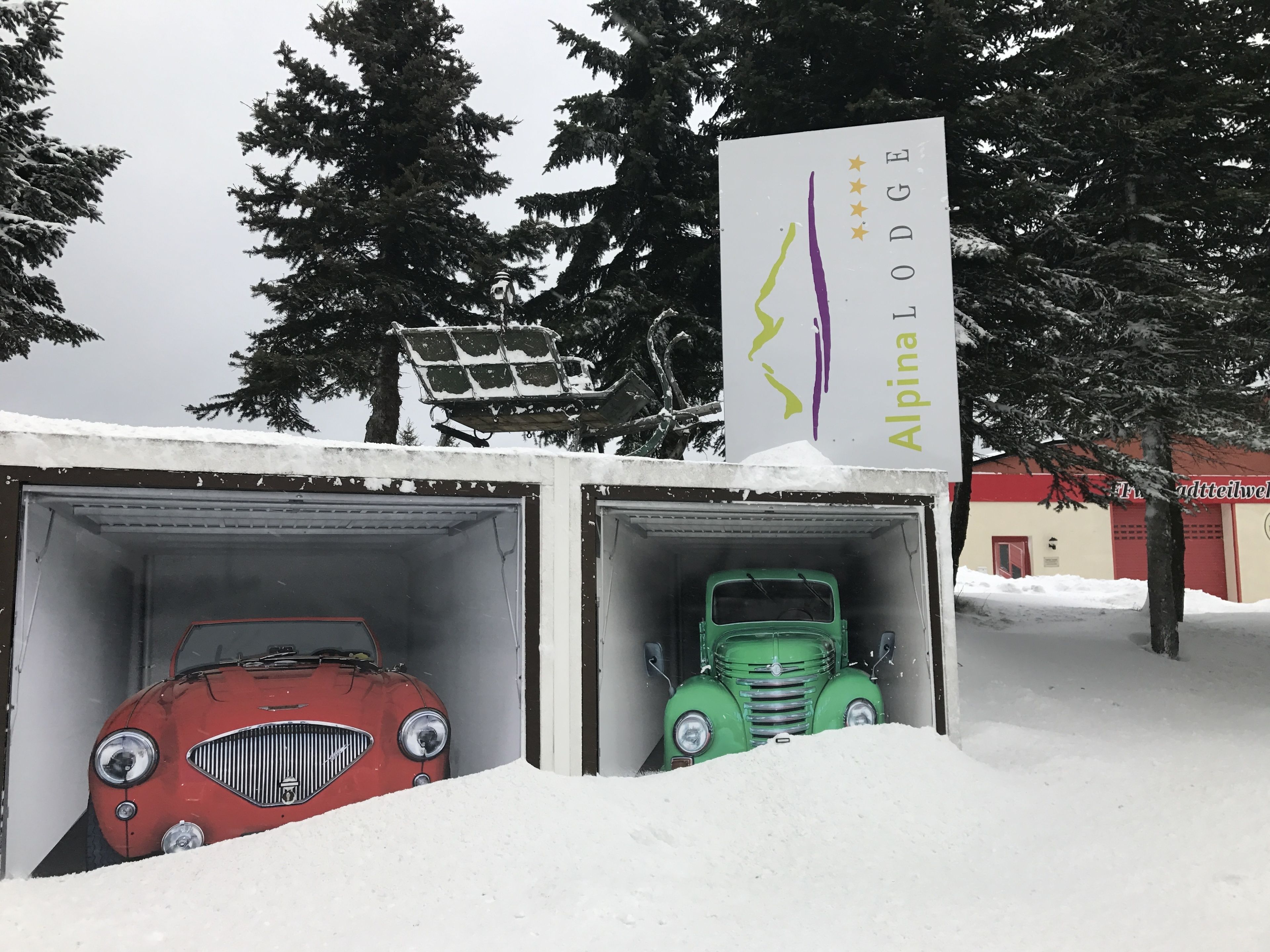 Two vintage cars parked in snow-covered garages with a sign for 'Alpina Lodge' in the background.