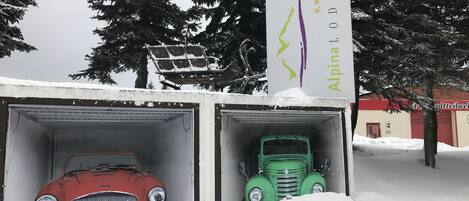 Two vintage cars parked in snow-covered garages with a sign for 'Alpina Lodge' in the background.