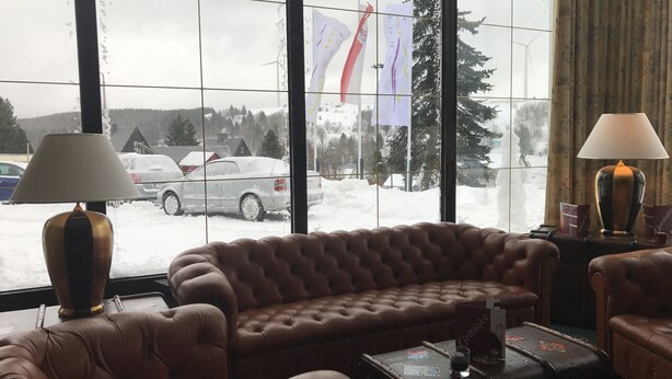 Ein gemütliches Zimmer mit einer braunen Ledersofa, zwei Lampen, einem kleinen Tisch und einem Fenster mit Blick auf eine verschneite Landschaft und Flaggen.