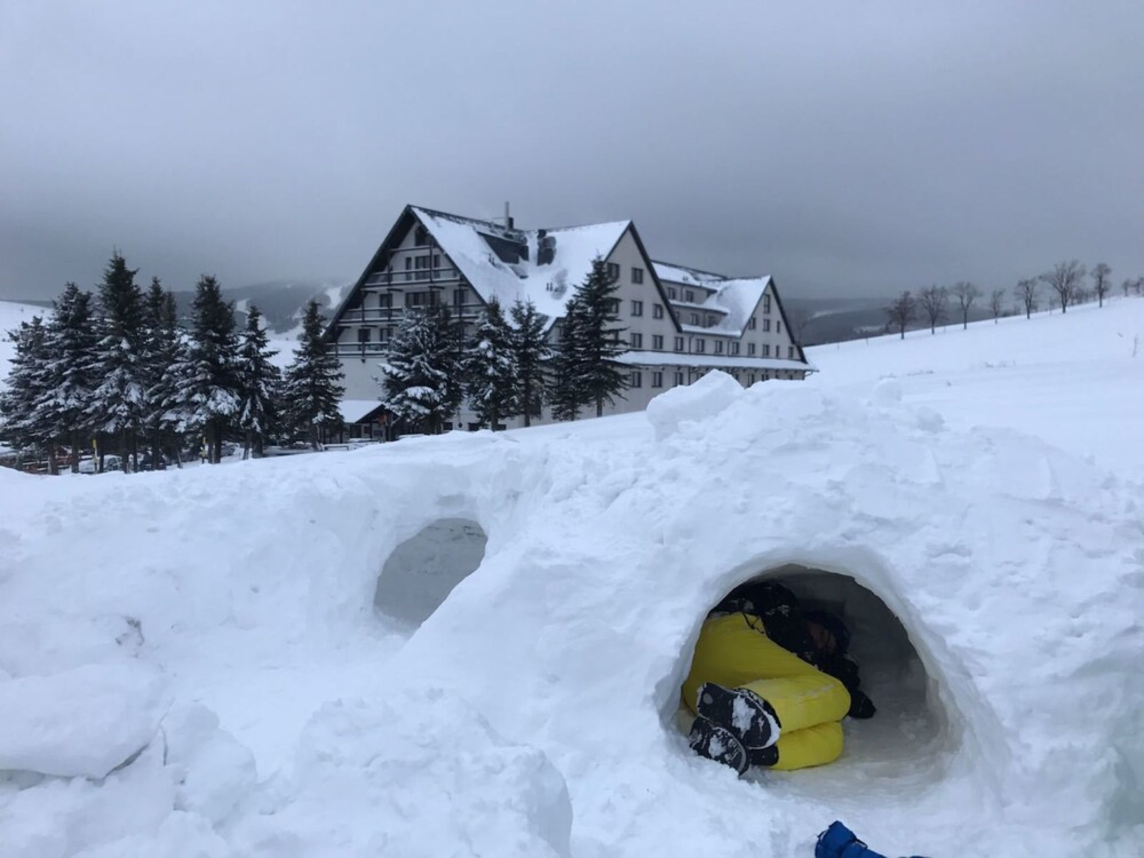 A snow-covered landscape with a large building in the background and a snow tunnel in the foreground.