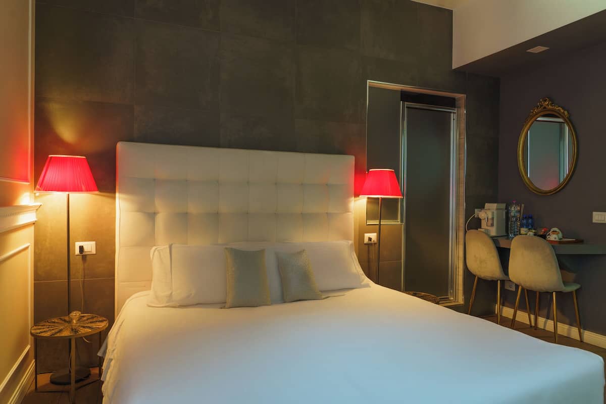 A neatly made bed with two pillows, a bedside table with a lamp, a mirror, and two chairs in a hotel room.