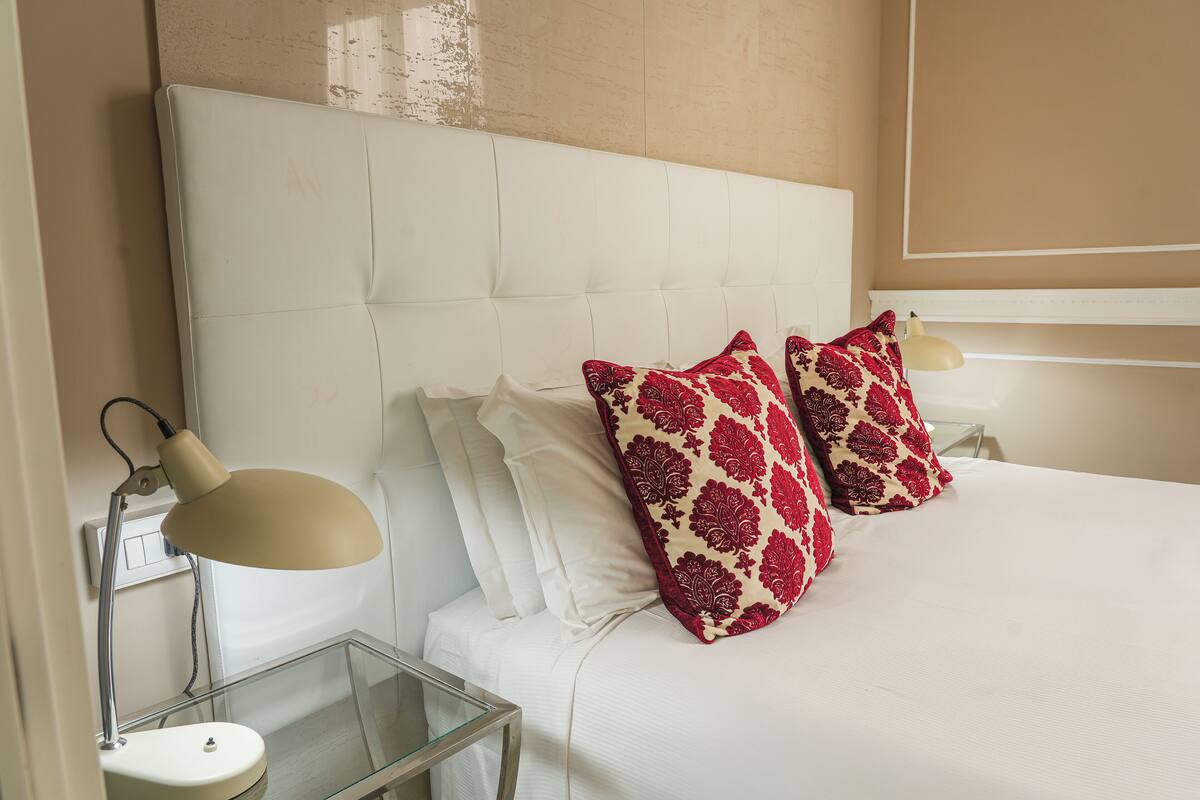 A neatly made bed with white bedding and two decorative pillows, a bedside lamp, and a glass side table.
