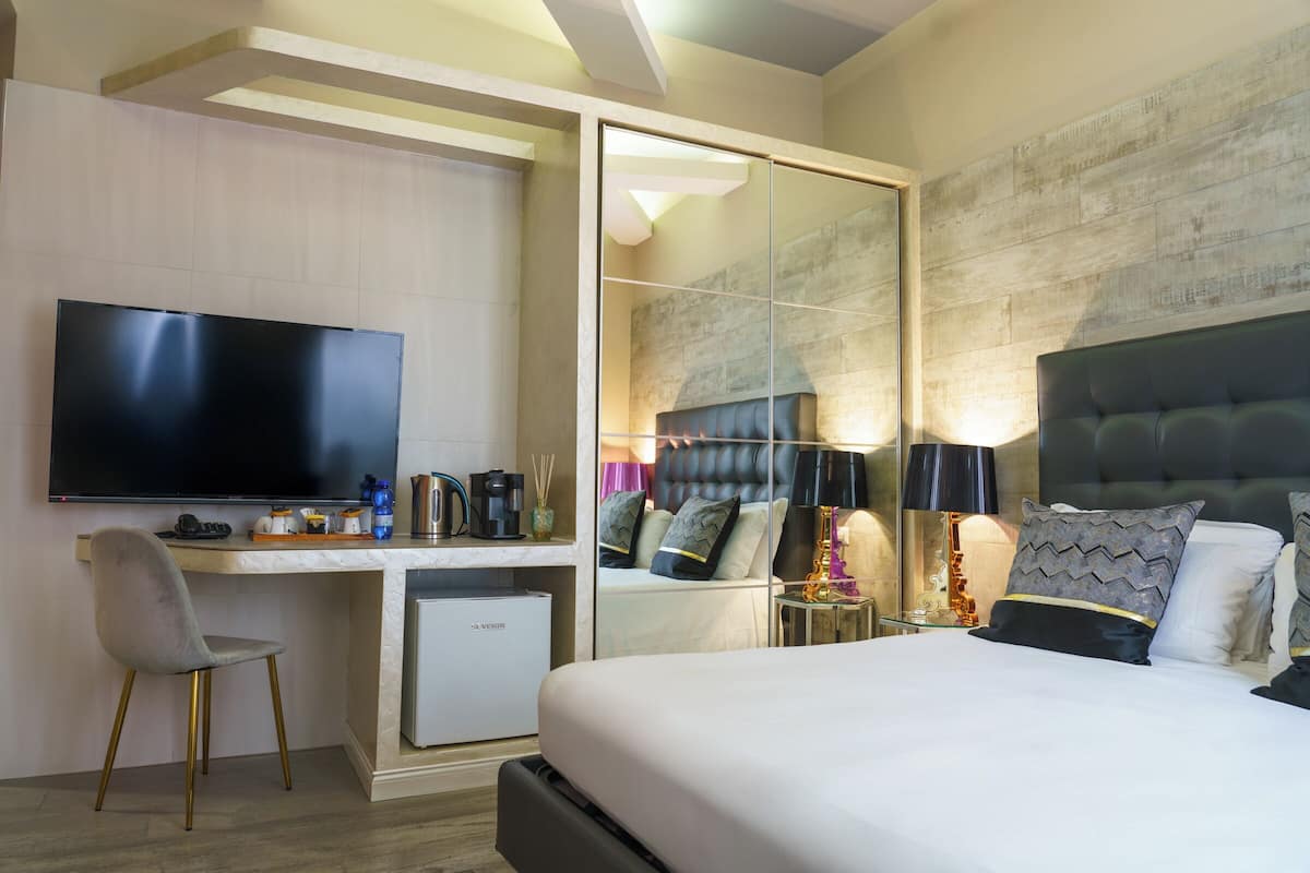 A modern hotel room with a large flat-screen TV, a comfortable bed with multiple pillows, a desk with a computer, and a small refrigerator.