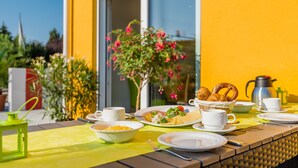 Une table de petit-déjeuner dressée avec des assiettes, des tasses et une cafetière, sur fond d’un mur jaune et d’une fenêtre.