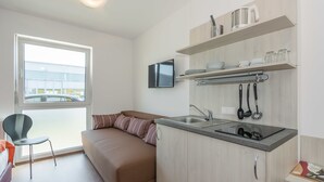 Un espace de vie compact comprenant un coin cuisine, un canapé, un fauteuil et un téléviseur fixé au mur.