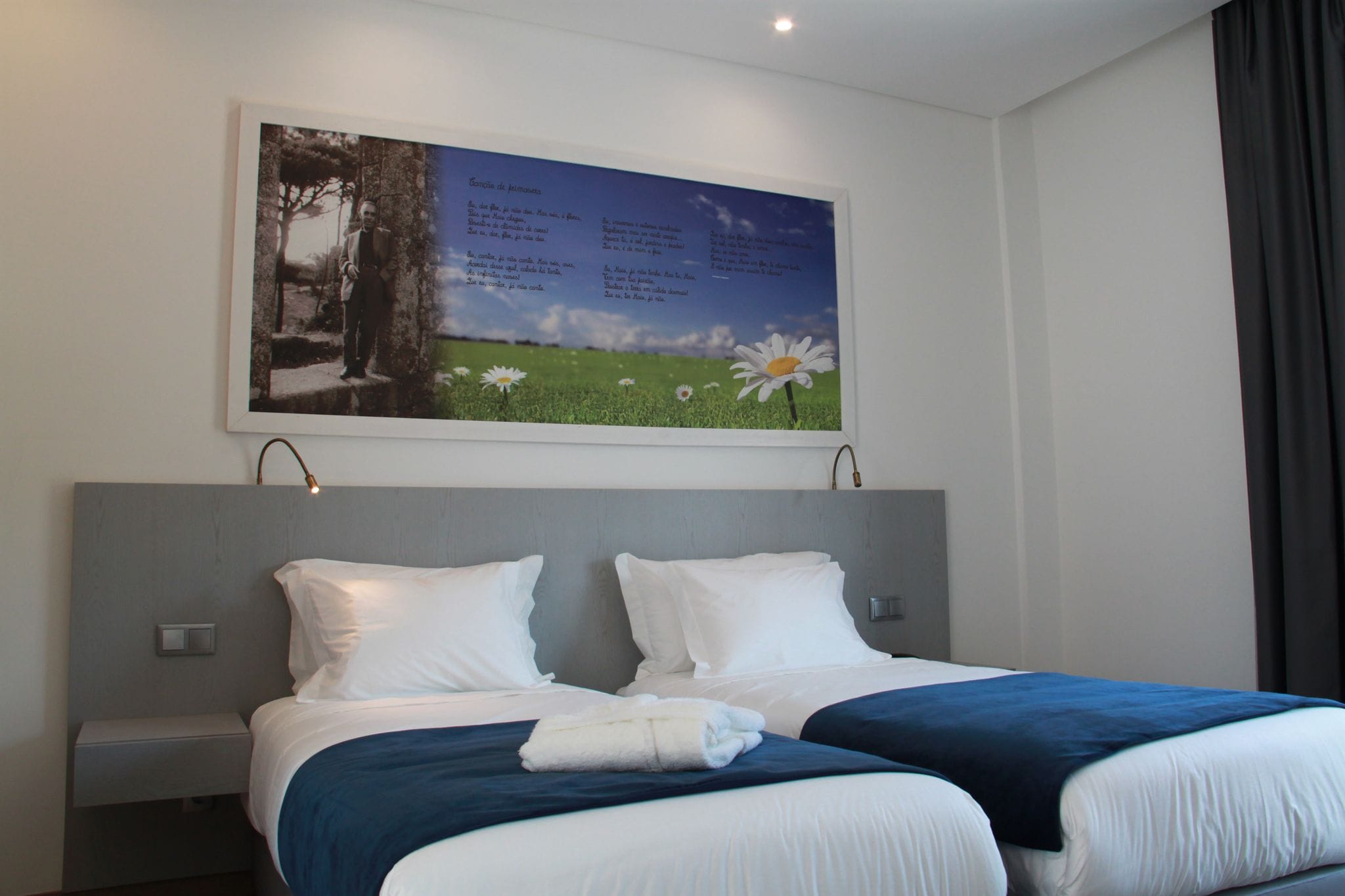 A hotel room with two beds, a nightstand, a wall-mounted artwork, and a window with curtains.