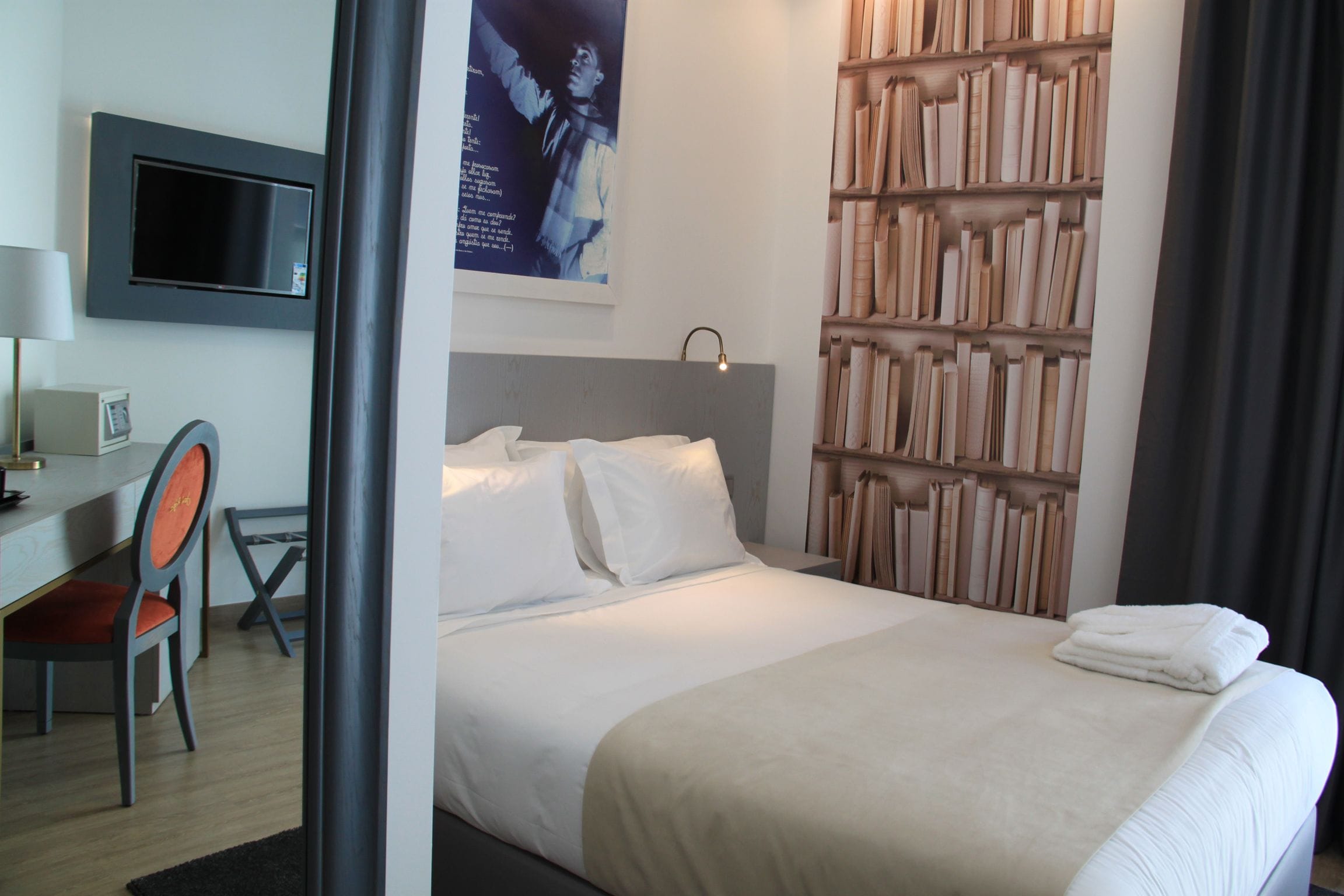 Ein Hotelzimmer mit Bett, Schreibtisch, Stuhl, Bücherregal und Flachbildfernseher.