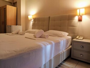 Una cama bien hecha con sábanas blancas y toallas enrolladas, una mesita de noche con lámpara y teléfono, y un armario de madera al fondo.