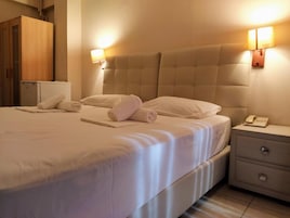 Una cama bien hecha con sábanas blancas y toallas enrolladas, una mesita de noche con lámpara y teléfono, y un armario de madera al fondo.