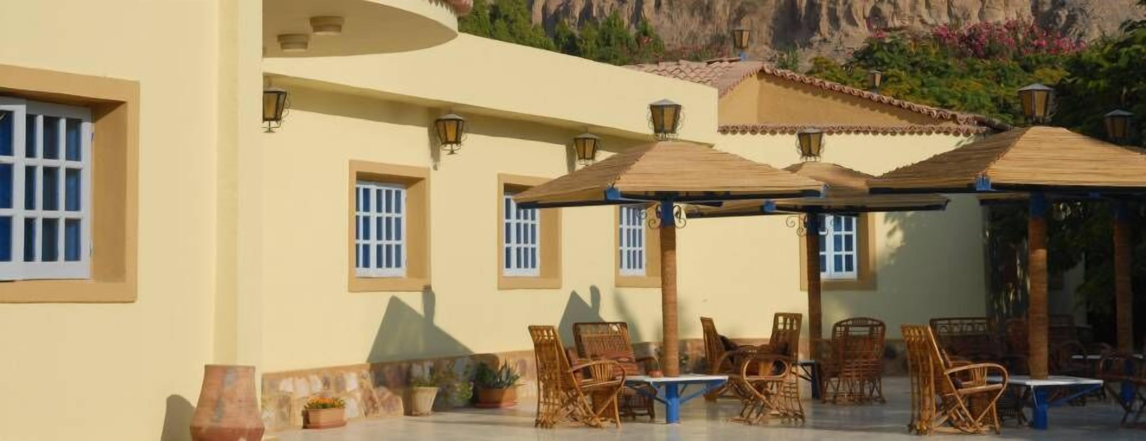 An outdoor patio area with wicker furniture and umbrellas, surrounded by a building with multiple windows and a tiled roof.