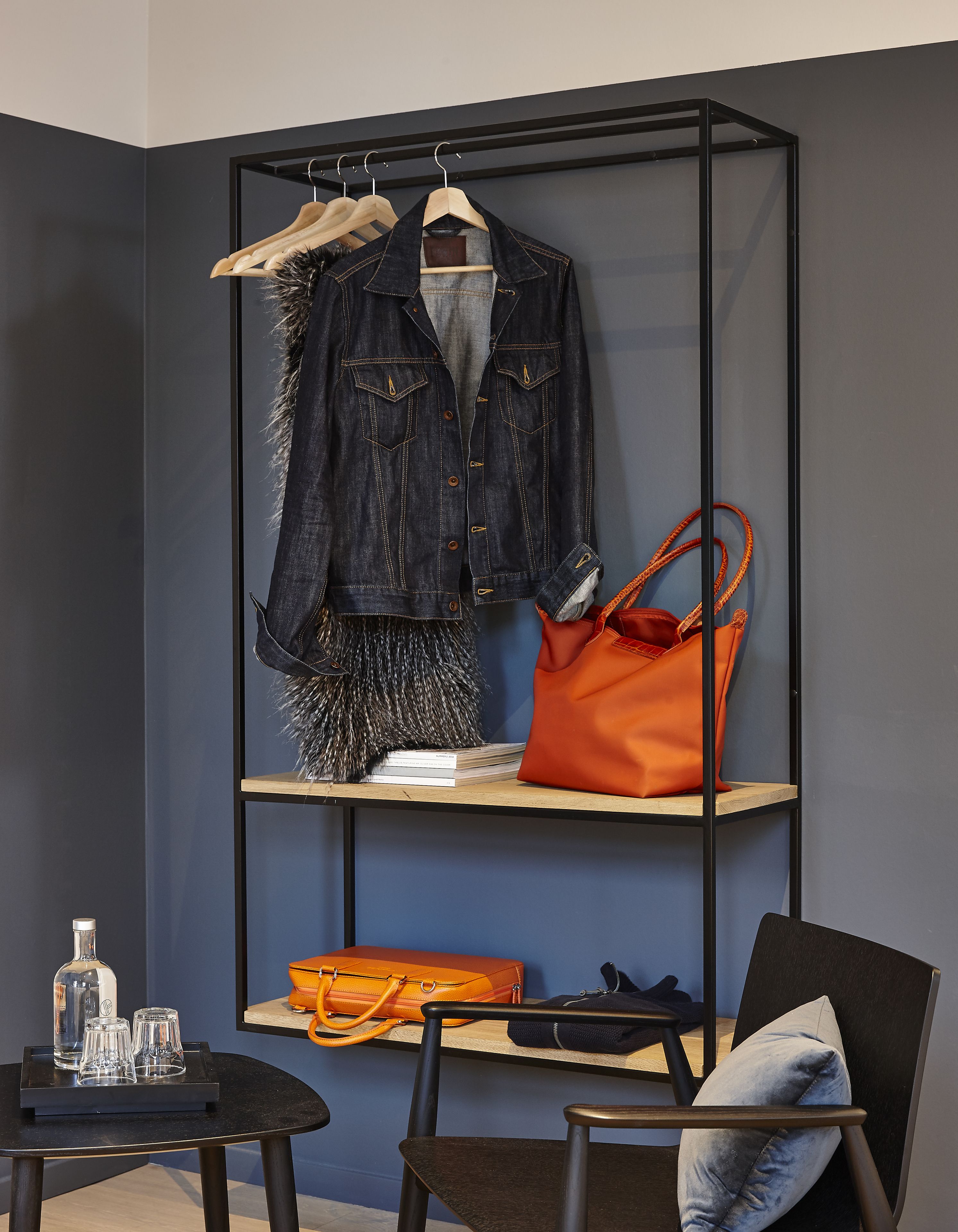 A room with a black metal shelving unit holding a denim jacket, a pair of shoes, and a handbag. Below the shelves, there is a small table with a glass bottle, a glass, and a black chair.