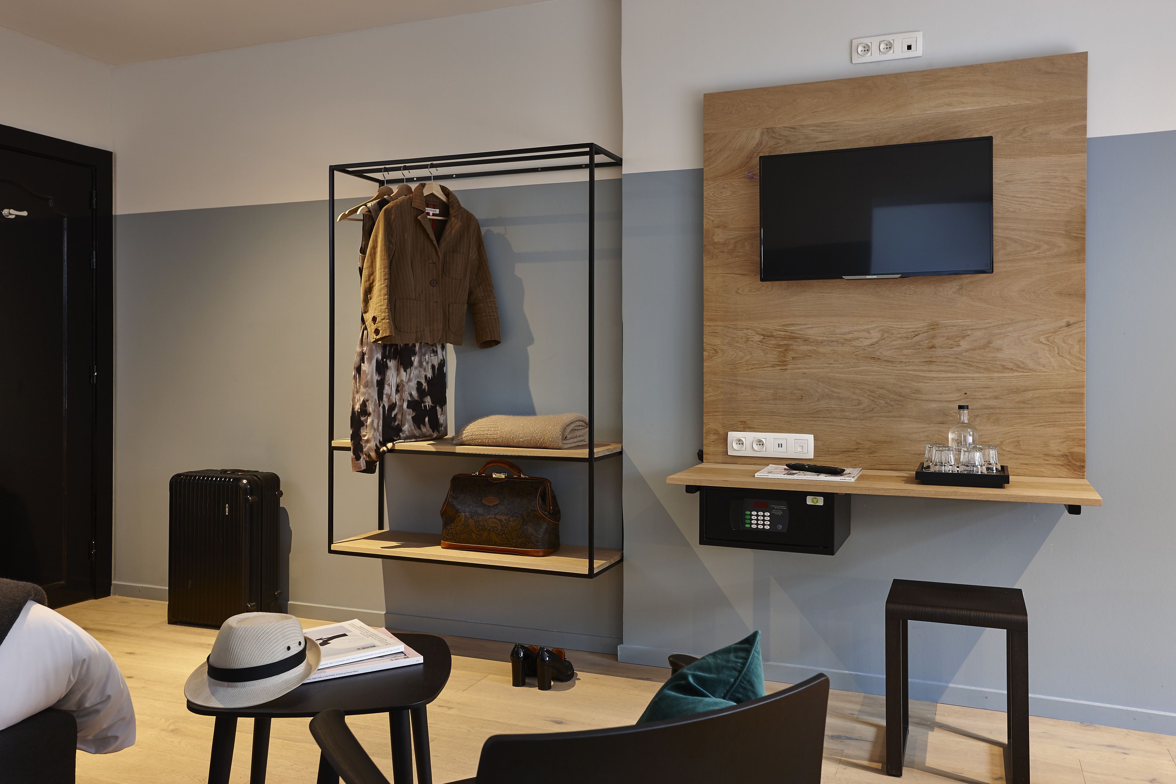 A modern hotel room with a wooden wall, a mounted TV, a shelf with a suitcase, a coat rack with clothes, a small table with a hat and glasses, and a chair.