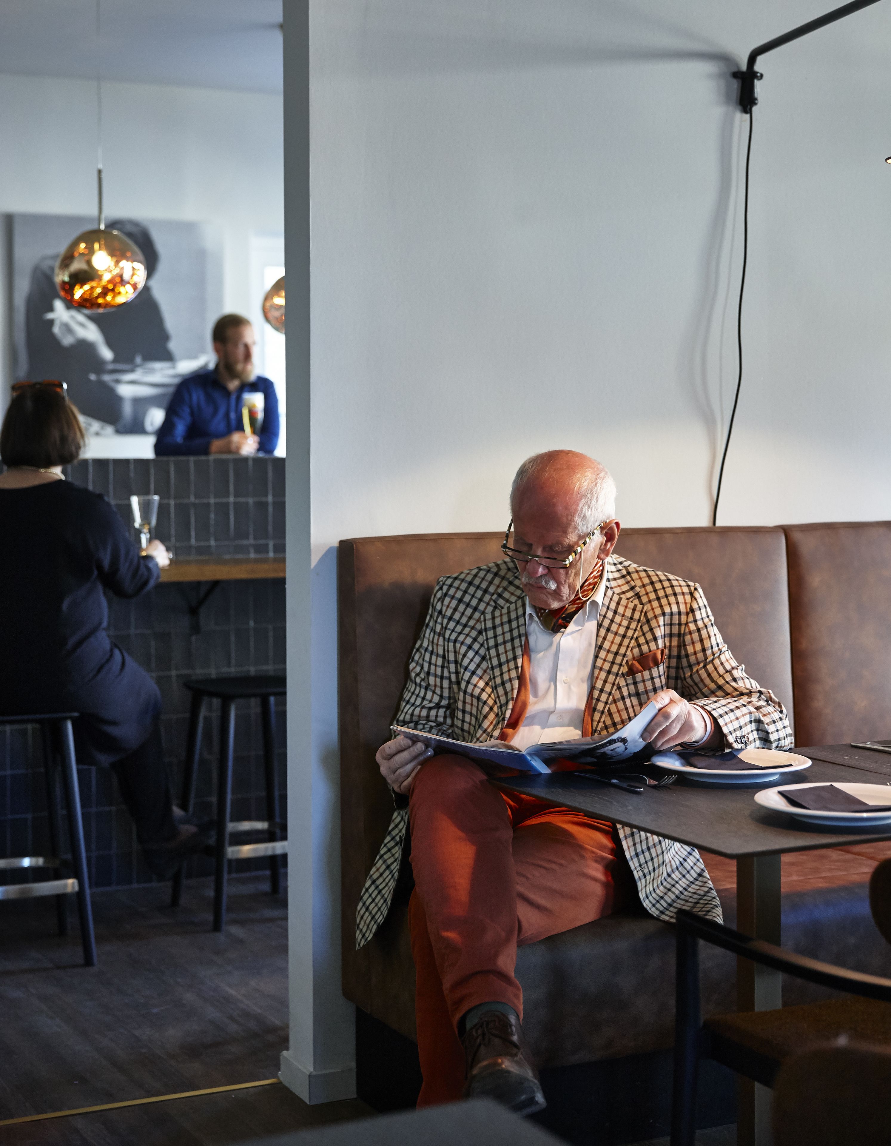 A man seated on a brown leather couch, reading a book. A woman is seated at a bar in the background.