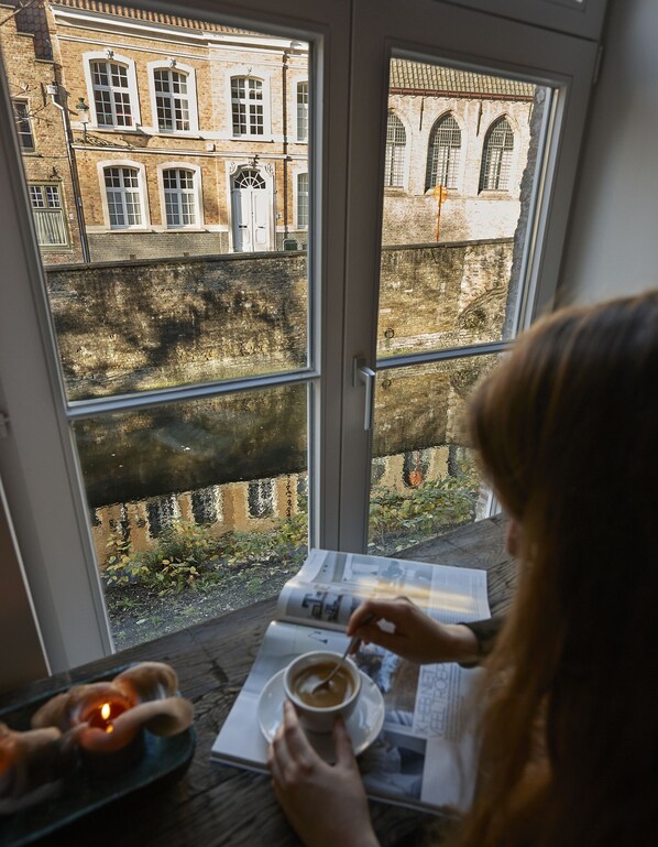 A person sitting at a table with a cup of coffee, reading a magazine, with a view of a brick building and a river outside a window. - Monsieur Ernest (Bruges)
