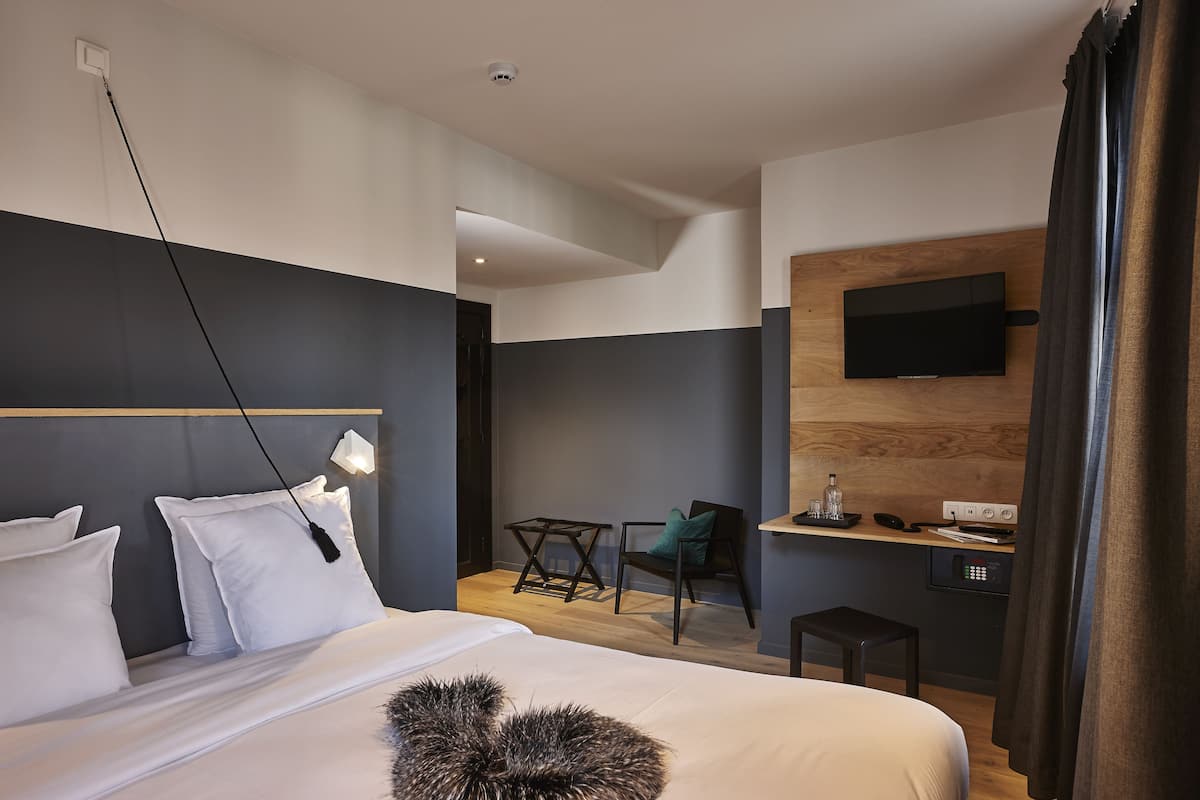 A modern hotel room with a large bed, a desk, a chair, a TV mounted on the wall, and a small table with a bottle and glasses.