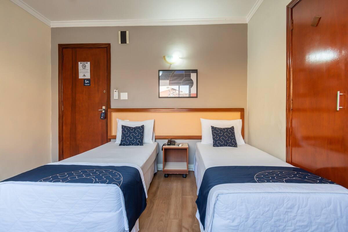 A hotel room with two single beds, a nightstand, a wall-mounted light, and a door with a sign.