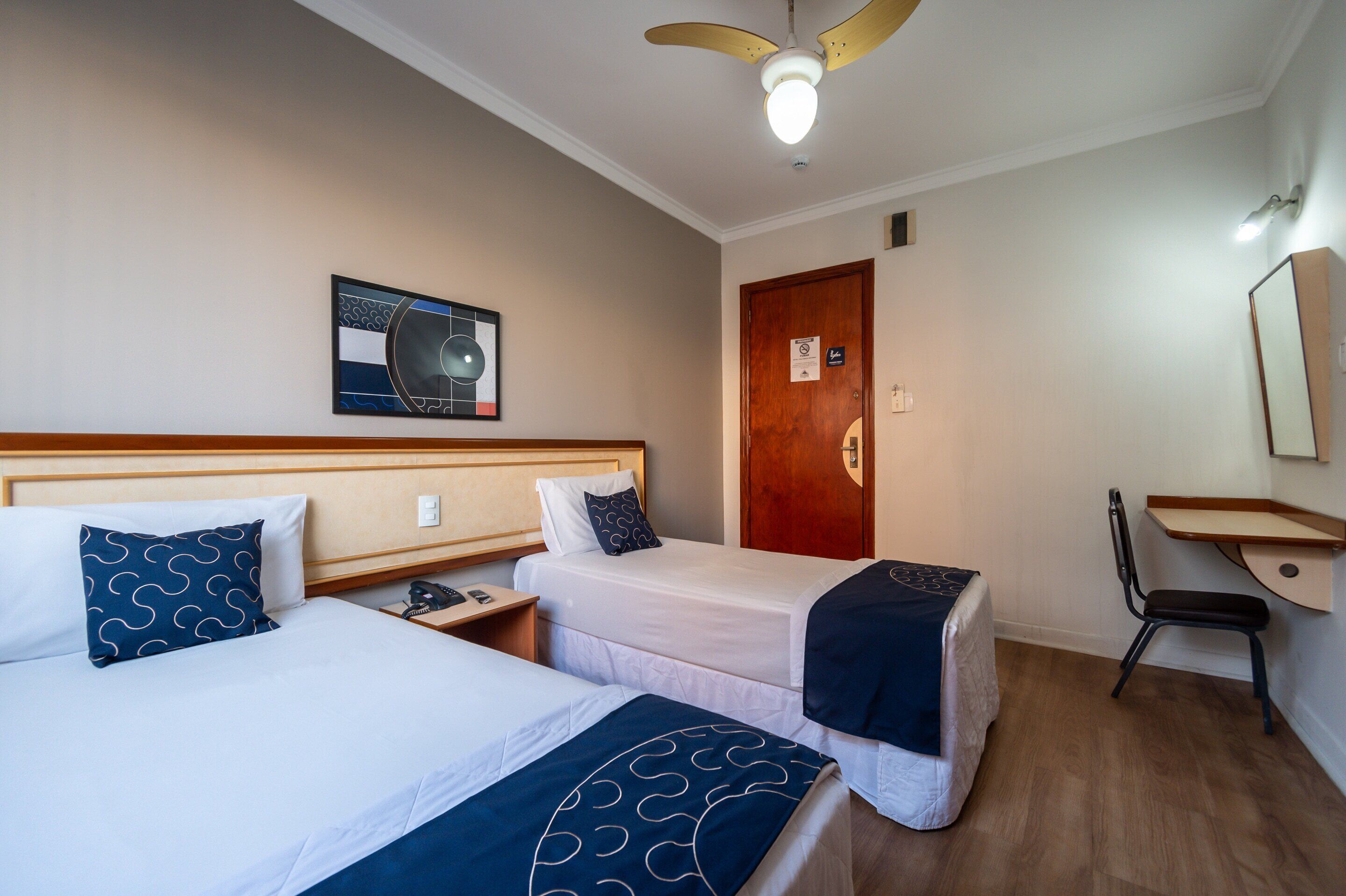 A hotel room with two single beds, a desk, a chair, a ceiling fan, and a wall-mounted artwork.