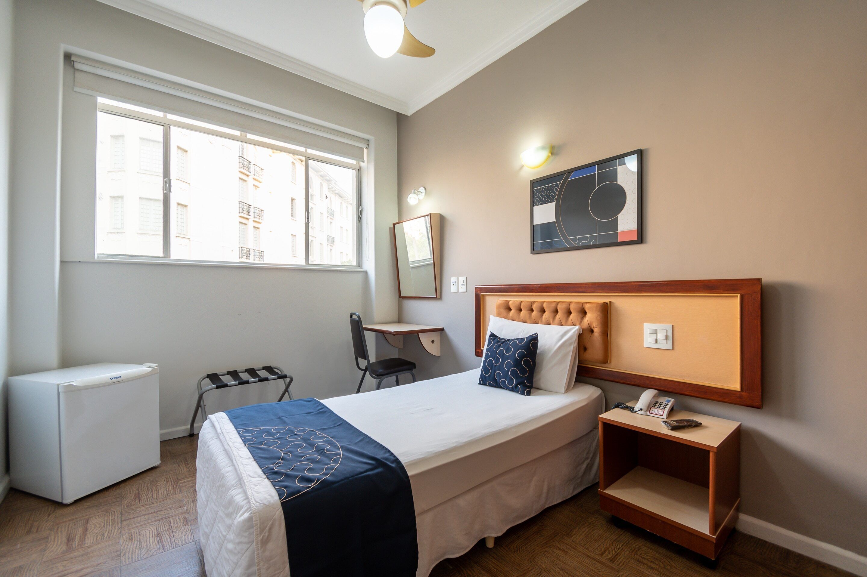 A hotel room with a bed, a desk, a chair, a small table with a phone, a heater, and a window with a view of buildings.