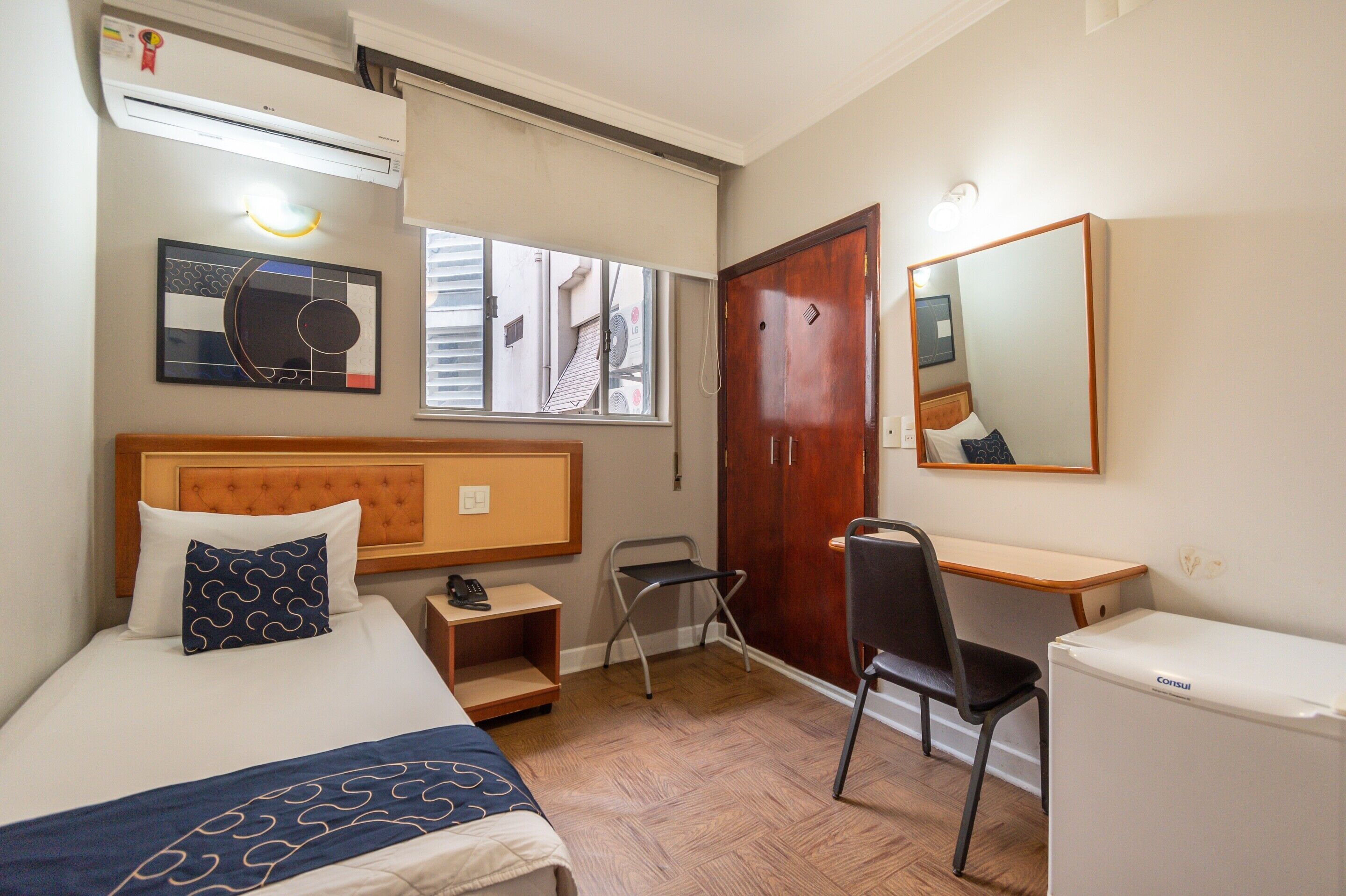 A hotel room with a bed, bedside table, chair, desk, and a refrigerator.