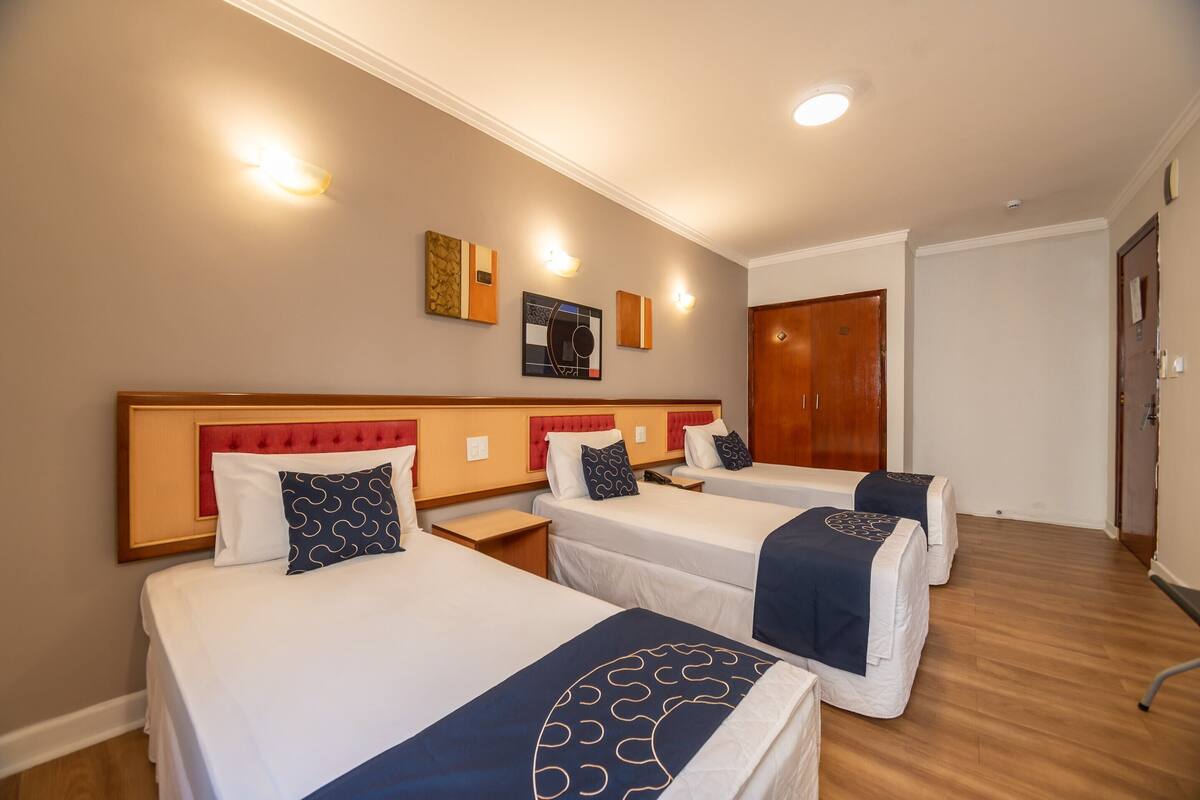 A hotel room with three single beds, a wooden headboard, bedside tables, and wall-mounted light fixtures.