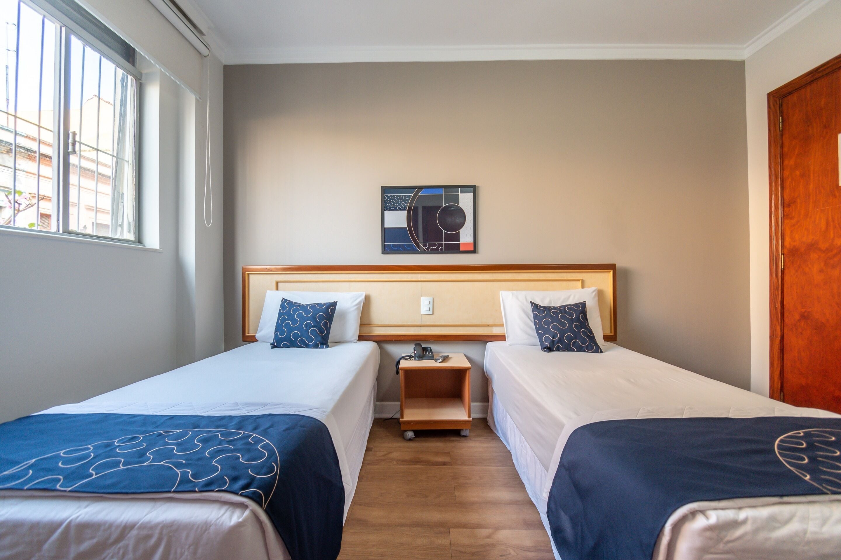 A hotel room with two single beds, a wooden nightstand, a wall-mounted artwork, and a window with bars.