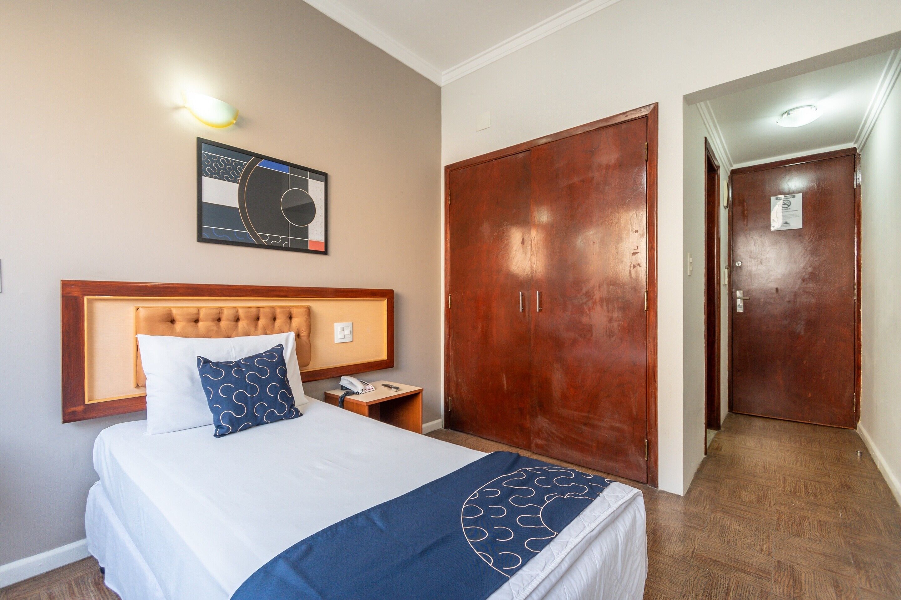 A hotel room with a bed, a wooden wardrobe, a nightstand, and a wall-mounted artwork.
