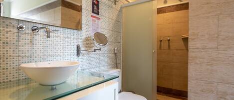 Un baño moderno con un lavabo de vidrio, armarios blancos, un inodoro y zona de ducha con mampara de cristal.