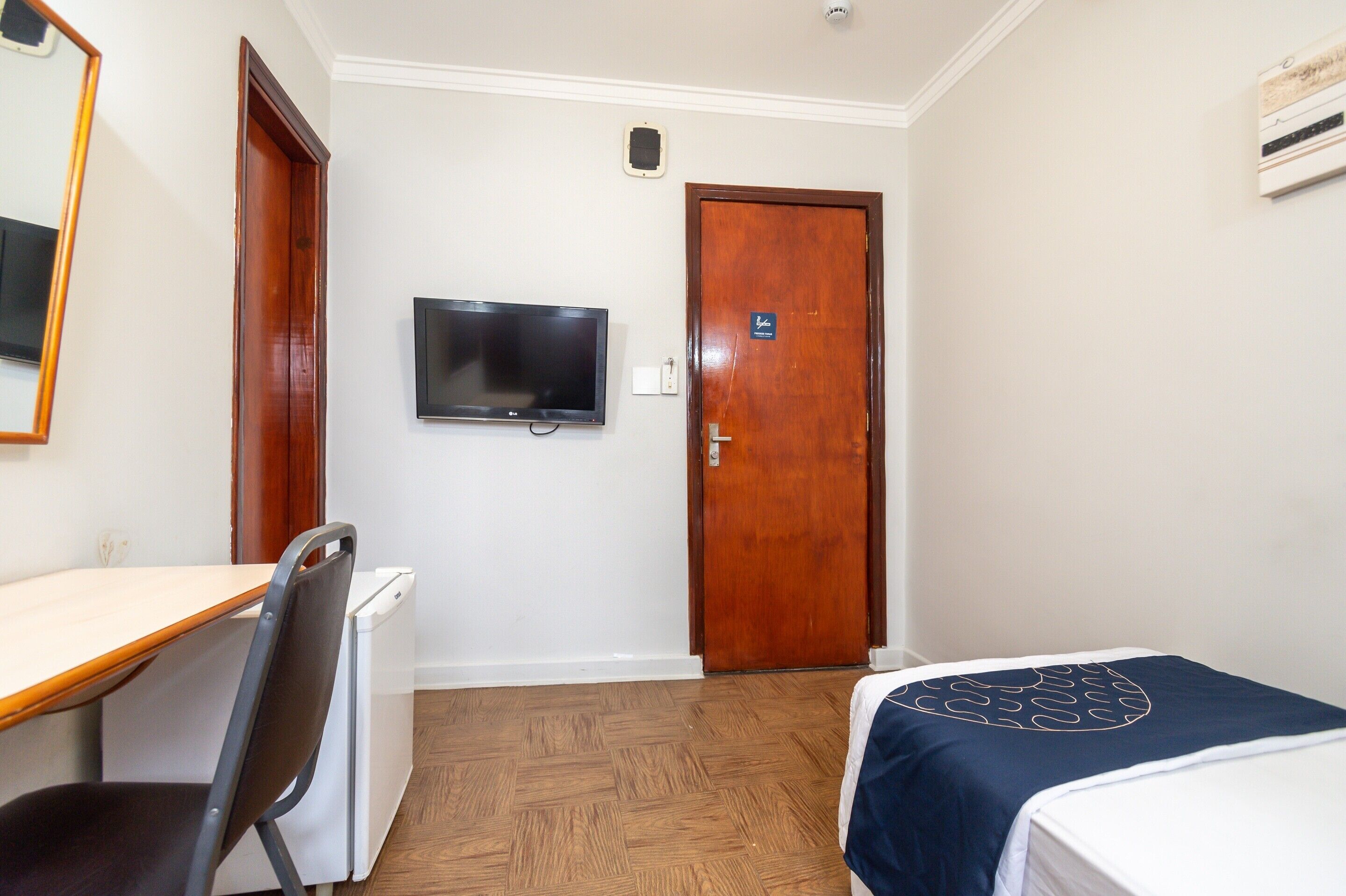 Habitació d'hotel amb escriptori de fusta, televisió penjada a la paret, llit amb edredó blau marí i porta de fusta.
