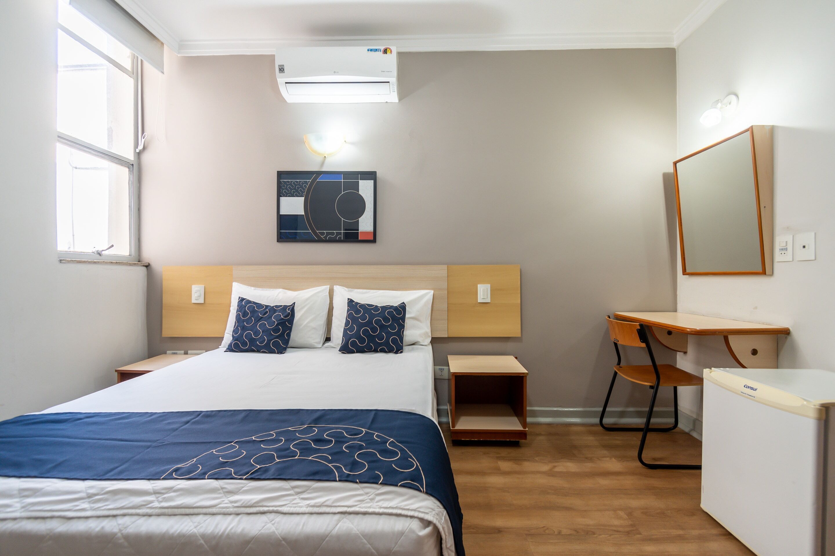 A hotel room with a bed, a desk, a chair, a small table, an air conditioner, a wall-mounted light, and a window.