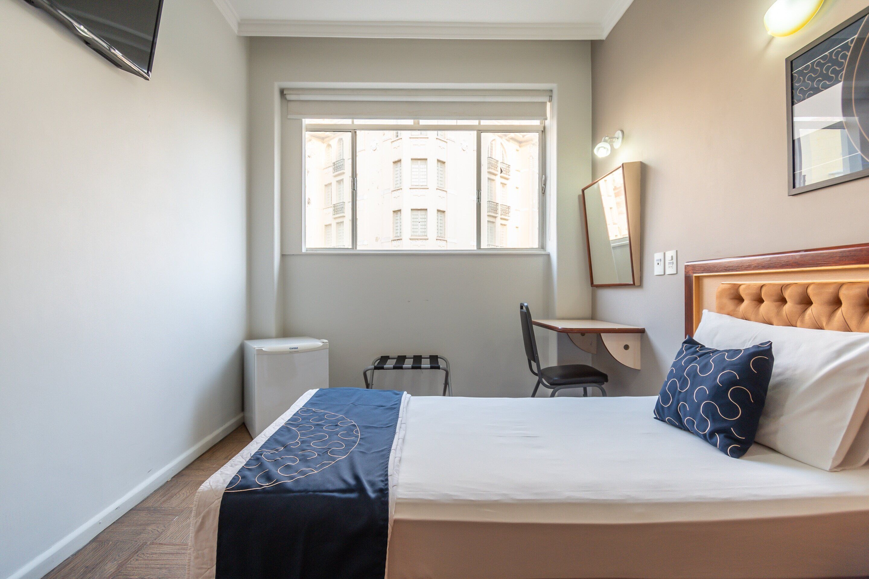 A hotel room with a bed, a desk, a chair, a window with a view of buildings, and a wall-mounted TV.