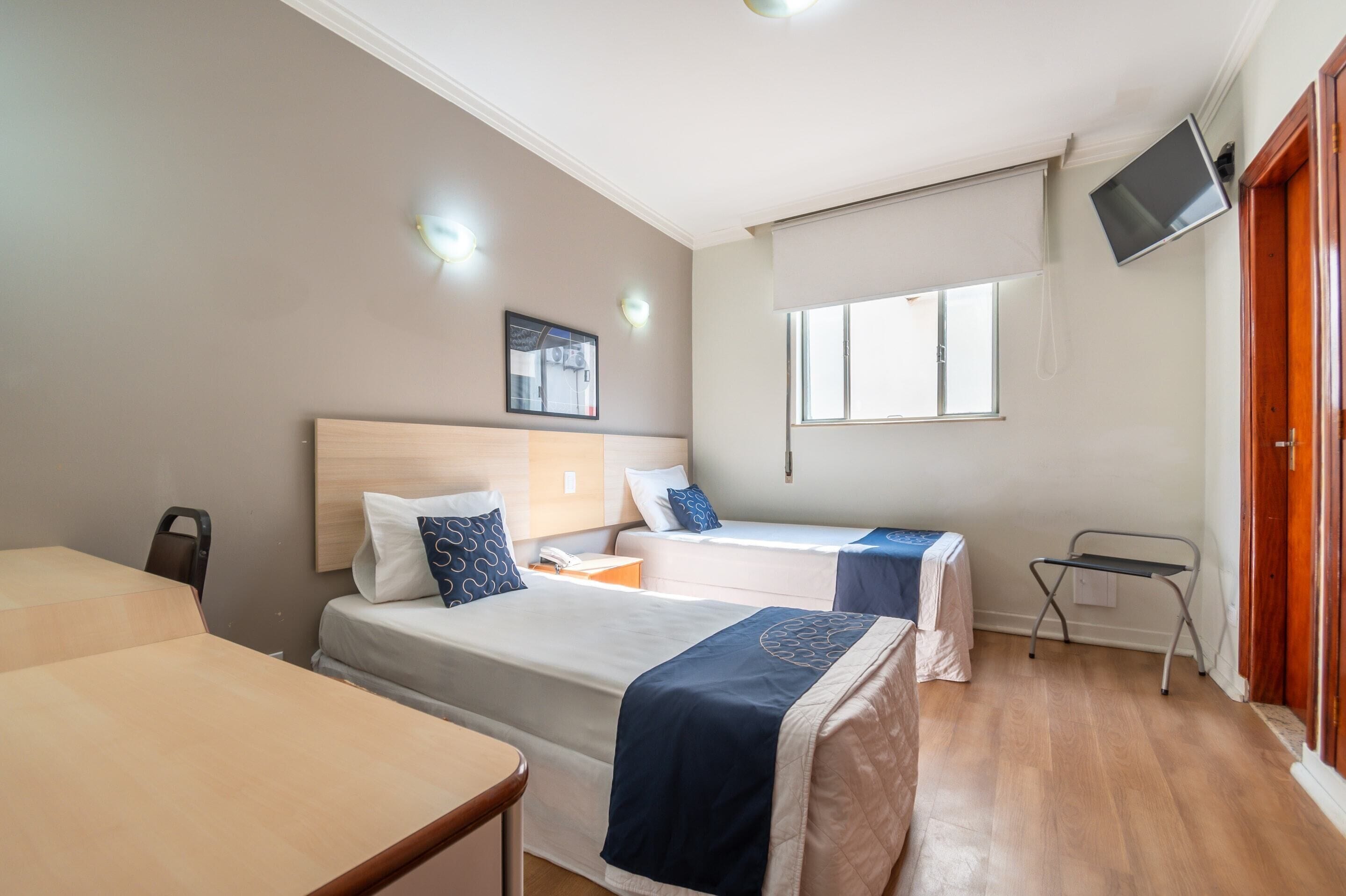 Une chambre d’hôtel avec deux lits, un bureau, une chaise, un téléviseur fixé au mur et une fenêtre.