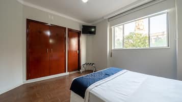 Quarto com cama, guarda-roupa, televisão e janela com vista para um edifício.