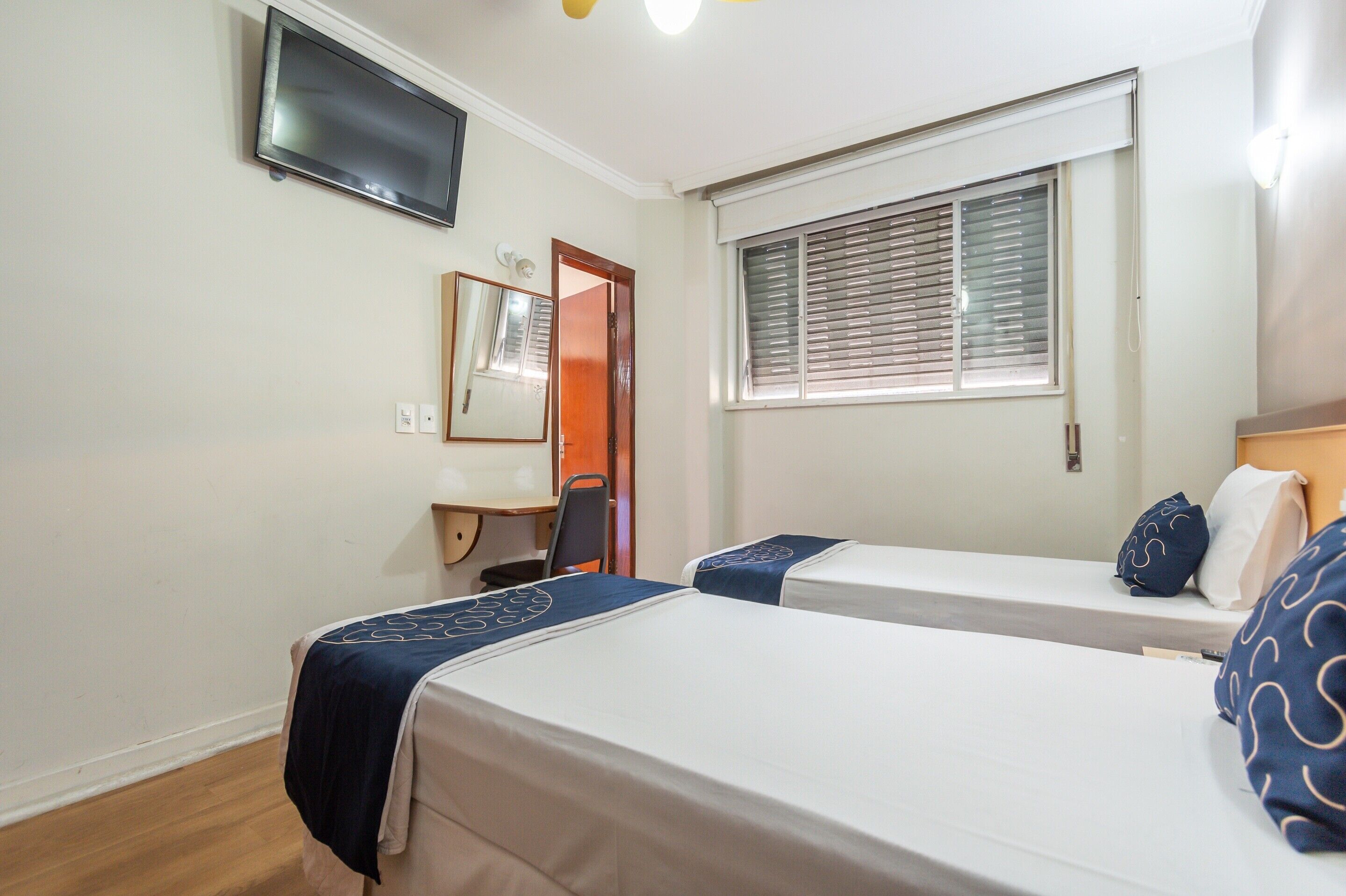 Une chambre d’hôtel avec deux lits, un bureau, une chaise, une télévision et une fenêtre avec des stores.