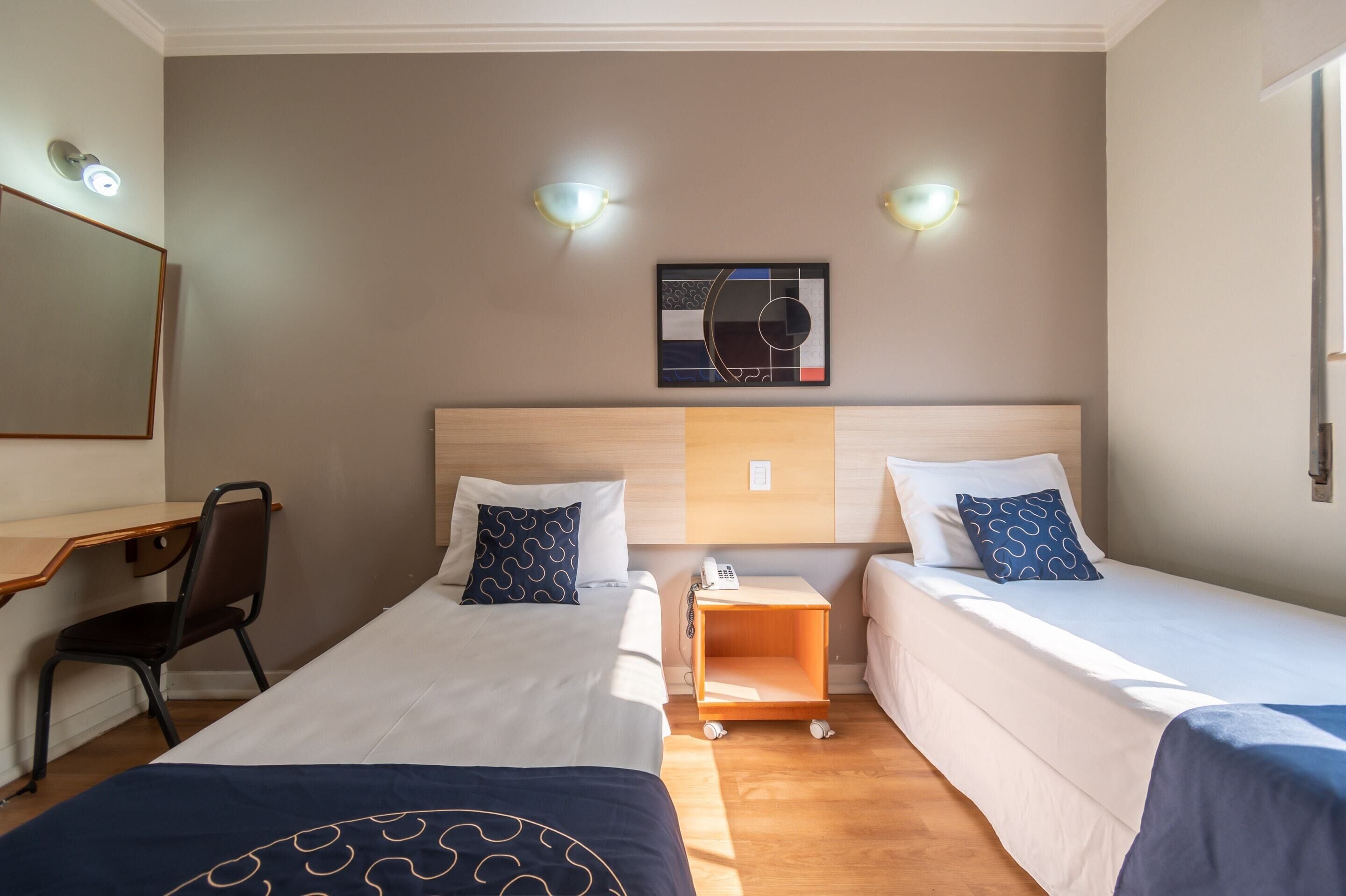 Ett hotellrum med två sängar, ett skrivbord och en stol. Det finns väggmonterade lampor och en inramad tavla ovanför sängarna.
