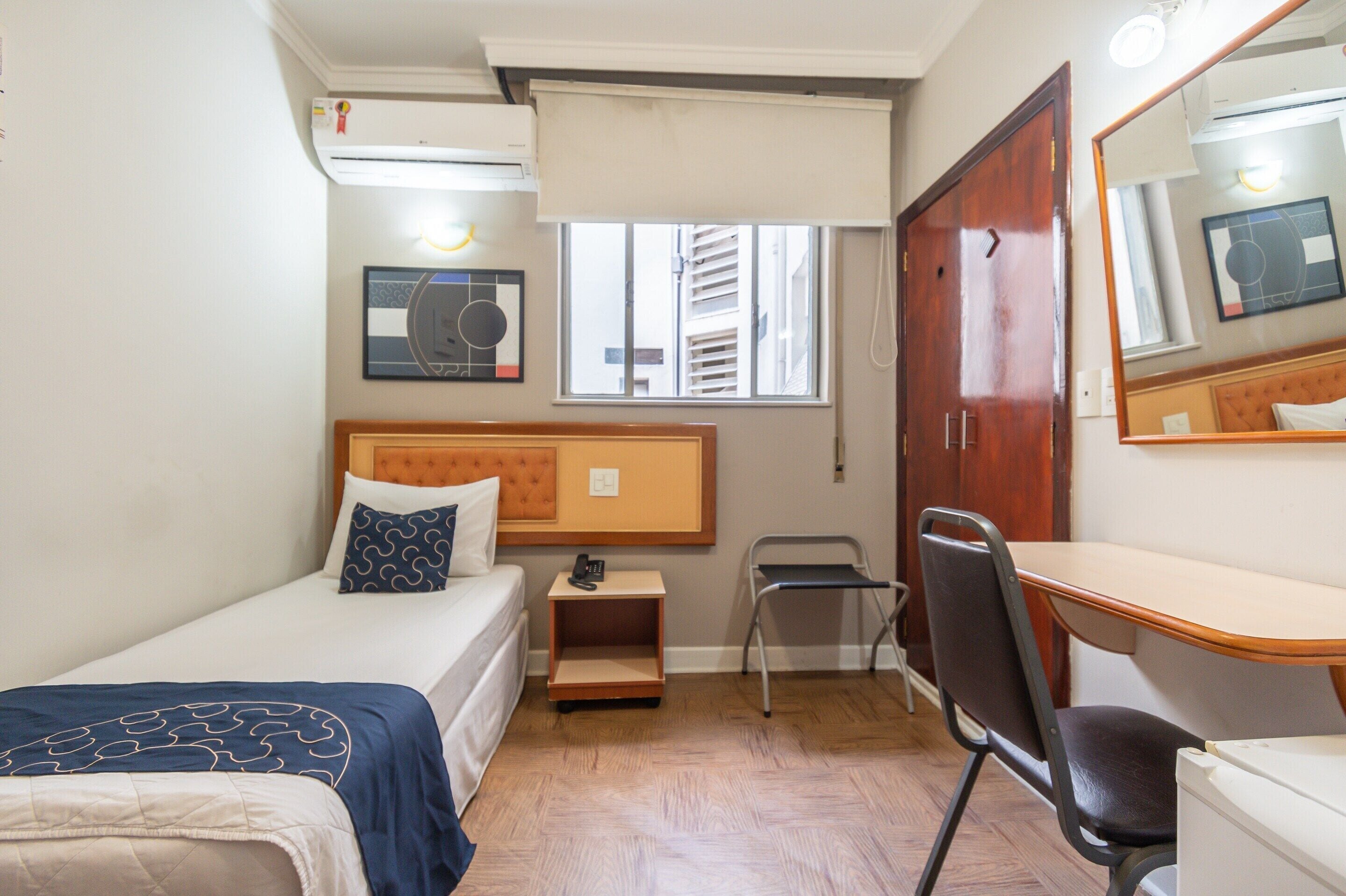 A small hotel room with a single bed, a desk, a chair, a wardrobe, and a window with blinds.