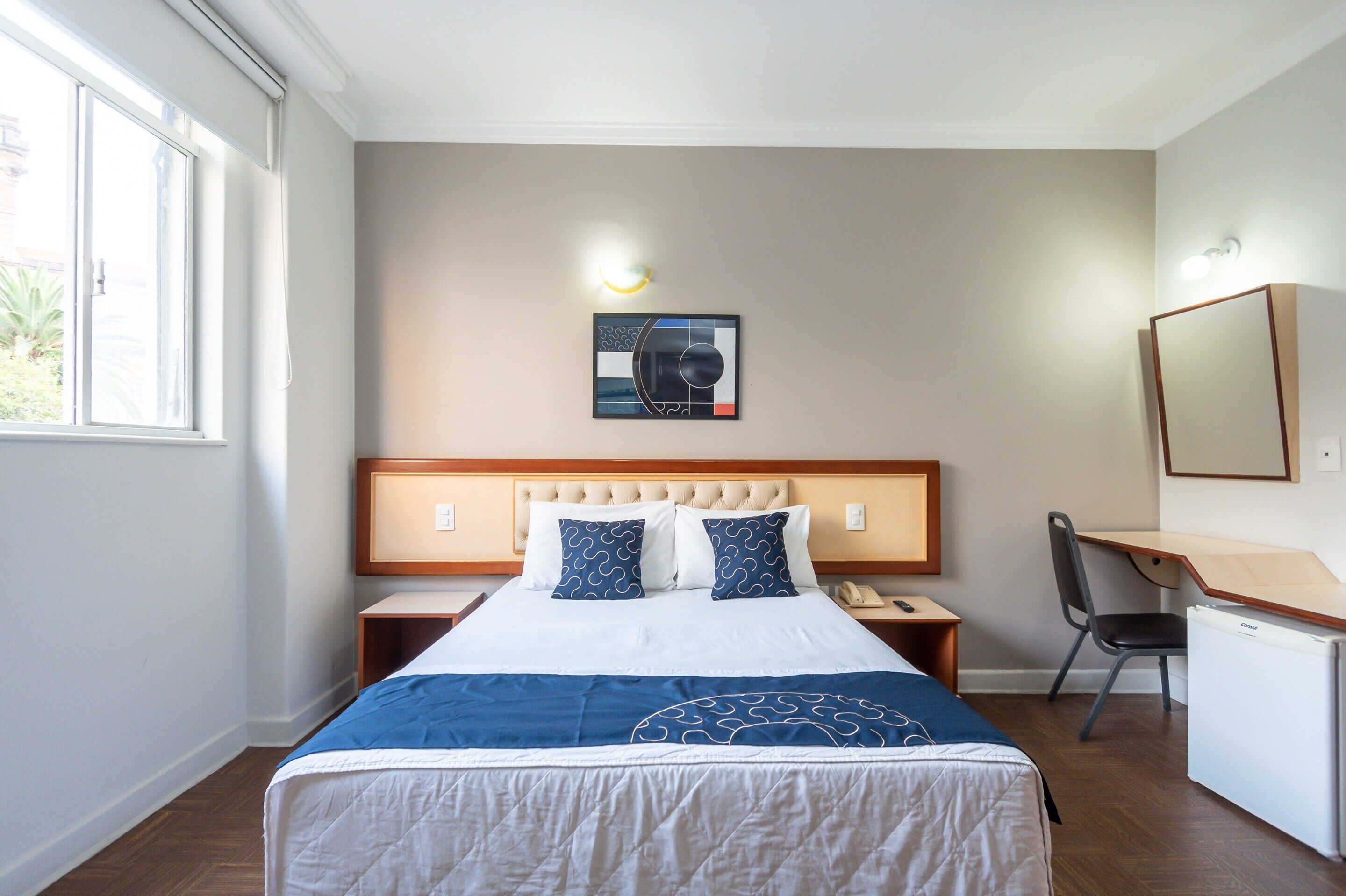 A hotel room with a bed, a desk, a chair, a small refrigerator, and a wall-mounted artwork.