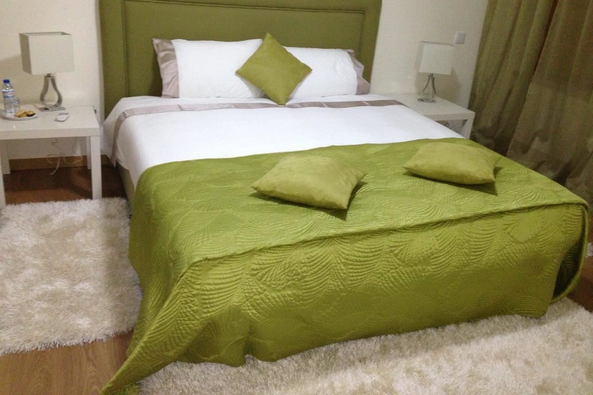 Ein ordentlich gemachtes Bett mit grüner Bettwäsche, weißen Kissen und einem grünen Zierkissen. Zwei Nachttische mit Lampen und ein flauschiger, weißer Teppich sind ebenfalls vorhanden.