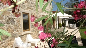 A stone building with a window and a door, outdoor seating area with white chairs and a table, and pink flowers in the foreground. - Otium Country House (Ansiao)