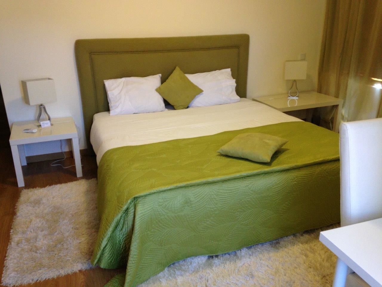A neatly made bed with a green bedspread and pillows, a white bedside table with a lamp, and a white chair.