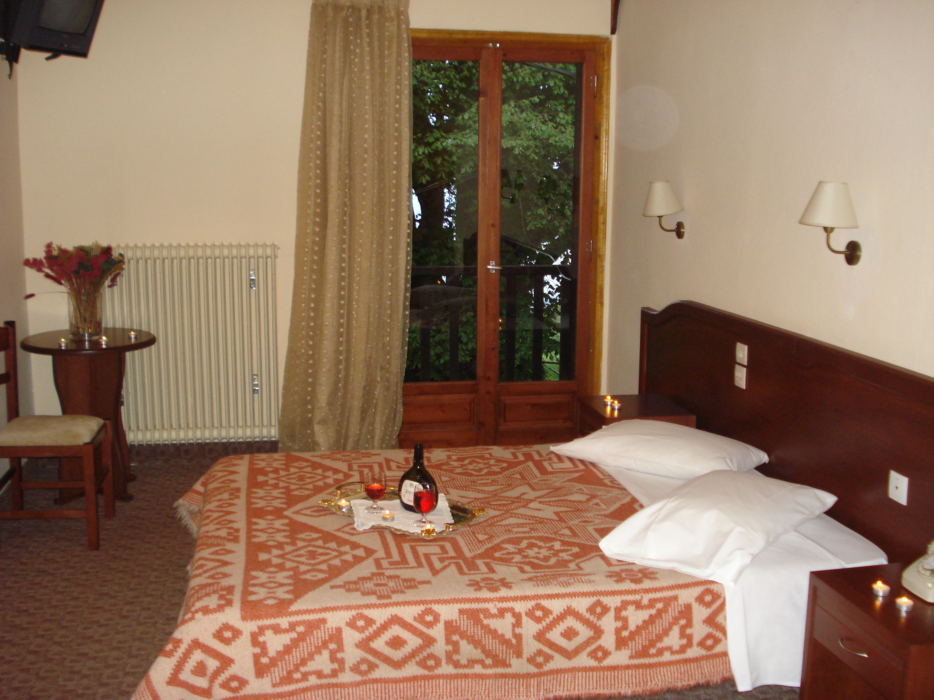 A hotel room with a bed, a chair, a table with a vase of flowers, a window with curtains, and a view of trees outside.